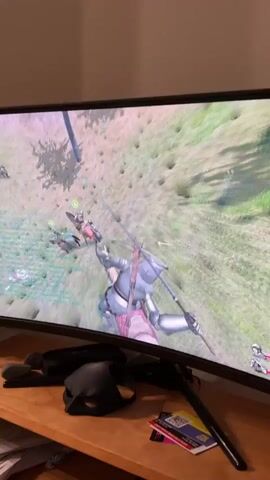 The video shows a wide-screen computer monitor displaying a video game with a medieval battle scene. The player character, a knight in armor, is in the foreground, wielding a sword and shield. Numerous other soldiers and horses are visible on a grassy, sloped battlefield in the background, engaged in combat. To the left of the monitor is a black mask, and to the right are several cards and a pen. In the lower portion of the screen, a person's hands are on a keyboard. A man with brown hair and a mustache is partially visible in the right-hand side of the frame, looking towards the screen. The overall environment appears to be a room with warm lighting.