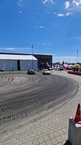 The video shows a car drifting around a paved track under a bright blue sky with scattered clouds. There are two cars visible in the frame, one yellow and one orange. The yellow car is a hatchback and the orange car appears to be a sedan. The track is marked with cones and has visible tire marks from previous drifts. In the background, there is a large white tent-like structure and a darker building with an arched entrance. Several other tents and vehicles are visible further in the background. The camera is positioned at a low angle, looking up towards the action. The overall scene depicts an outdoor motorsports event, likely a drifting competition or demonstration.