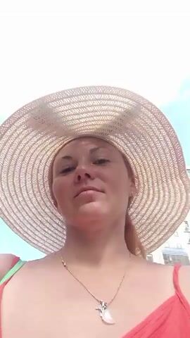 The video shows a close-up of a woman wearing a wide-brimmed straw hat and a red dress. The camera is positioned below her face, looking upwards. She has a necklace with a pendant. The background consists of a bright blue sky with some clouds and the side of a building. The woman's face is partially in shadow due to the hat. The video appears to be a selfie-style recording.