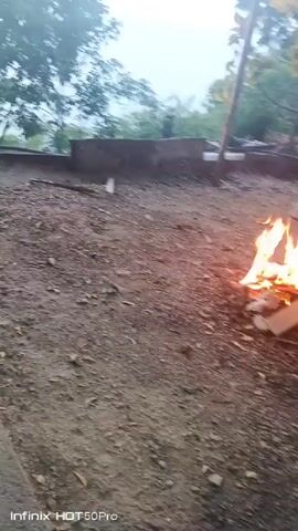 A small bonfire burns in a dirt clearing. The fire is made of sticks and some white material, possibly cloth or paper. The ground is covered with dirt, small rocks, and dry leaves. In the background, out of focus, are some trees and what appears to be part of a wooden structure. A person's legs and feet, wearing light-colored pants and dark shoes, are visible on the right side of the frame, near the fire. Another person is partially visible in the upper right corner. The lighting suggests it is dusk or early morning.