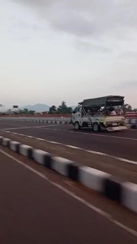 The video shows a view from a vehicle moving on a highway. The camera is pointed forward and slightly to the right, capturing the road, median, and vehicles in the distance. The sky is overcast and appears to be dusk or dawn. In the middle lane of the opposite direction, a small white truck with a covered bed is visible, carrying several passengers. To the left of the truck, a motorcycle is also traveling in the same direction. The median between the lanes is lined with white and black barriers. The road surface is asphalt. The overall impression is of a typical highway scene during low light conditions, with traffic moving at a moderate pace.