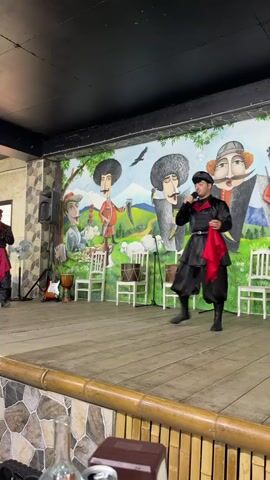 The video shows two men performing on a wooden stage in front of a colorful mural. Both men are wearing traditional black outfits with red sashes. The man in the center is singing into a microphone and holding a red cloth. The man on the left is standing in a dynamic pose. In the background, there are white chairs, musical instruments, and a fan. The mural depicts stylized figures in a landscape setting. There is also a table in the foreground with bottles and what appears to be a menu.