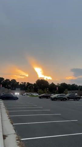 The video shows a parking lot with several cars parked. The sky is filled with dramatic clouds at sunset, with rays of sunlight breaking through. The sun appears to be setting behind a line of trees in the background. The parking lot is paved and has white lines marking the parking spaces. The overall impression is a serene and beautiful natural scene.