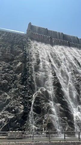 A large man-made waterfall cascades down a tiered concrete or rock structure under a clear blue sky. Water flows over a series of vertical columns at the top, then streams down a wide, textured surface, creating multiple rivulets and sheets of white water. A metal railing is visible at the bottom of the frame, separating the viewer from the falling water. No people or other significant objects are present.