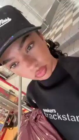 A person is holding a camera looking up at their face, wearing a black baseball cap with a green bill, gold hoop earrings, and a black long-sleeved shirt with "Seekers trackstar" printed on it. They are looking directly at the camera with a slight pout. They are holding a large, maroon plastic bag in front of them. In the background, there is a rack of items, including what appears to be a magazine and some greenery. The floor has a gray and white striped pattern.