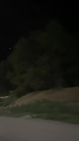 A low-light video captures a nighttime scene from a moving vehicle. The camera pans from right to left, revealing a dark, tree-lined embankment in the foreground. A dirt road or unpaved area runs along the bottom of the frame. In the background, a two-story building with visible windows is partially obscured by trees. Further in the distance, lights suggest a more populated area or roadside establishments. A bright, distant light, possibly a star or a light fixture, is visible in the upper left quadrant of the screen. The overall atmosphere is dark and quiet, suggesting a rural or suburban setting at night.