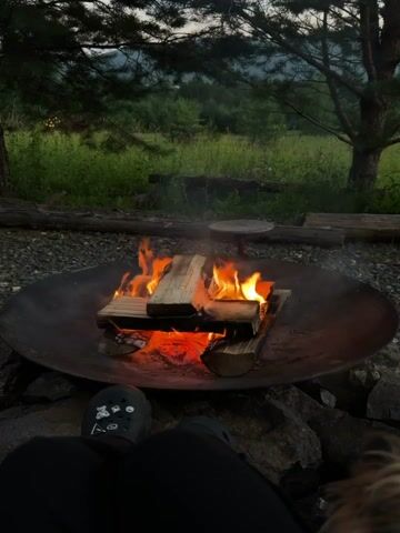The video shows a close-up view of a fire pit with a campfire burning. The camera is positioned low, looking up at the fire. In the foreground, a person's legs and feet are visible, wearing black pants and patterned Crocs. The fire is in a large, round, dark metal fire pit. There are several pieces of wood burning, with bright orange flames and glowing red embers. The background shows a grassy field with trees and a hint of mountains in the distance. The lighting suggests it is either early morning or late evening, with a soft, diffused light. A small portion of a dog's head and fur is visible on the right side of the frame. The overall scene is peaceful and suggests an outdoor setting, possibly camping or a backyard.
