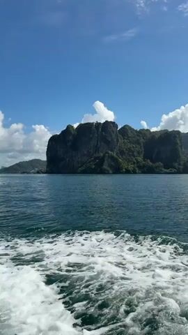 A boat is moving through the water, creating a wake. The water is blue and choppy. In the distance, there are large, rocky islands covered in green vegetation. The sky is blue with white clouds. The sun is shining.