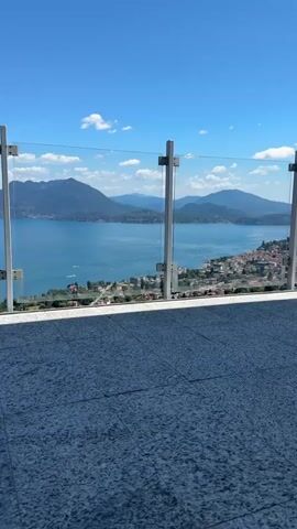 The video shows a panoramic view from a balcony or terrace overlooking a large body of water, likely a lake, with mountains in the distance. The foreground features a tiled floor and a modern glass railing. The lake is calm and blue, with several boats visible on its surface. Along the shoreline, a town or village with red-tiled roofs and scattered buildings is nestled among lush green trees. The sky is a clear, vibrant blue with a few scattered white clouds. The camera pans slowly across the landscape, offering a changing perspective of the scene. The overall impression is one of a beautiful, serene natural environment with a picturesque human settlement.