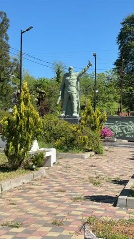 The video shows a park on a sunny day with a light-colored statue of a man in the center. The statue is of a man with a sword and his arm raised. Behind the statue is a stone wall with an image of soldiers on it. The park is decorated with green bushes and flowers. There is a bench to the left of the statue. The pavement is made of red and brown bricks. There are street lights on either side of the statue. A man is singing in the background.