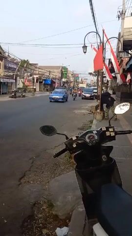 The video shows a street scene in what appears to be a town or city. On the right side of the screen, a black scooter is parked on the sidewalk. The street is paved and appears to have some cracks and unevenness. Several vehicles are present on the street, including a blue station wagon, a red scooter with two riders, a white truck, and other motorcycles. People are riding the motorcycles and the scooter. Buildings line both sides of the street, with some appearing to be shops. Red and white flags are displayed on poles along the street, suggesting a national holiday or celebration. The sky is overcast, and the lighting suggests it is daytime. The overall impression is of a moderately busy street with typical urban traffic.