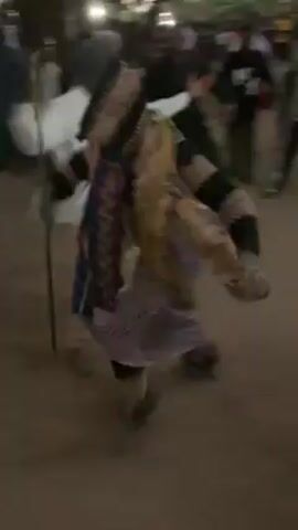 The video shows a person in a traditional costume dancing in a public space. The dancer is wearing a striped mask or head covering and is adorned with colorful fabrics and what appears to be straw or raffia fringes. The person is also holding a long, thin stick. A crowd of people surrounds the dancer, some of whom are clapping and recording with their phones. The setting looks like an outdoor gathering, possibly a village square, with a sandy or dirt ground. The light suggests it might be dusk. The mood of the scene appears celebratory and cultural.