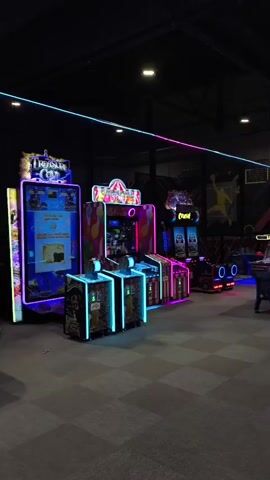 The video pans across an indoor arcade, showcasing various game machines illuminated by neon and LED lights. On the left, a game titled "Treasure Cove" is prominently displayed with a blue and green neon border. Adjacent to it are several upright arcade cabinets with colorful graphics and purple neon accents. Further to the right, a game called "Pogo Jump" features a blue platform and a vertical screen with cartoon characters. Another section of the arcade displays multiple racing-style games with glowing pink and blue trim, and what appear to be simulated vehicles for players. The floor is covered in a checkerboard pattern. The ceiling is dark, with thin neon strips of pink and blue light running across it. The overall ambiance is that of a dimly lit entertainment venue filled with vibrant, electronic game attractions. No people are visible in the footage.