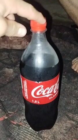 A hand with a light skin tone is opening a 1.5 liter bottle of Coca-Cola Classic. The bottle is mostly full of a dark brown liquid and is covered in condensation. The bottle has a red cap and a red label with white text. The background is dark and appears to be a blanket or similar fabric. A person's legs and feet are visible in the upper right corner of the frame.