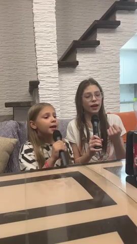 Two young girls are singing karaoke at home. They're sitting on a couch in front of a glossy brown table, each holding a microphone. The girl on the left has blonde hair and is wearing a striped shirt, while the girl on the right has long brown hair, glasses, and a white t-shirt. A phone or tablet is positioned on the table between them, presumably displaying the karaoke lyrics. Behind them, a staircase with wooden steps sits against a textured white wall. The couch has a patterned throw and a few pillows. The overall atmosphere is casual and fun.