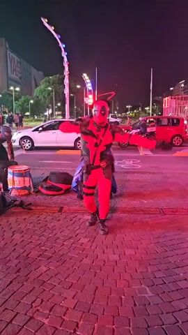 The video shows three people in costumes dancing in the street at night. One person is in a Deadpool costume, and the other two are in Spider-Man costumes with slightly different variations. They are performing in a public space with brick pavement, a busy street with cars, and buildings visible in the background. The lighting is a mix of streetlights and the colors of the city. The Deadpool character is central and performs dance moves with his arms outstretched. The Spider-Man characters move similarly, and one has a cowboy hat. There is a drum and some other items on the ground, suggesting they might be street performers.