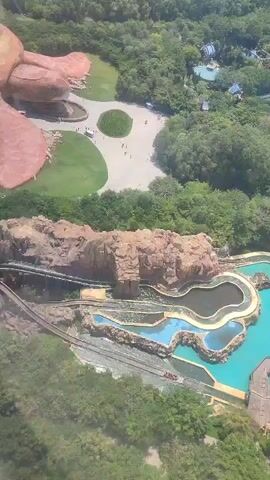 The video shows an aerial view of a theme park. A large, artificial turtle structure is prominent, lying on its stomach on a green lawn next to a paved area where people are walking. Below the turtle structure is a water ride featuring a series of pools and rocky terrain. A small log flume boat is seen on the ride. The scene is surrounded by lush greenery. In the background, there are buildings, a body of water, and what appears to be the ocean. The video is shot from an elevated perspective, possibly from a high building or aerial vehicle.