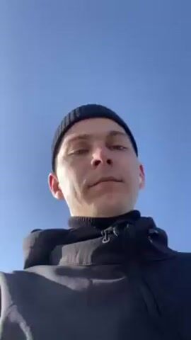 The video shows a man from a low angle against a clear blue sky. He wears a black beanie, a black turtleneck, and a black hooded jacket. The man has fair skin and short brown hair. He is looking directly at the camera. An earbud is visible in his right ear.