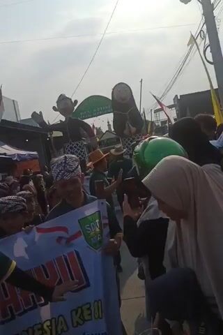 The video shows a parade with many people. In the foreground, a man in a green shirt and a traditional patterned headscarf is holding a banner. The banner has red and white writing on it, with a green and yellow logo. Behind him, other people are visible, some also wearing traditional headwear. In the background, there are large inflatable figures of a man and a woman dressed in traditional clothing. A sign is visible behind the inflatables with text that appears to be in Indonesian. There are also tents, flags, and other parade decorations. The sky is bright, suggesting it is daytime. The overall atmosphere seems festive.