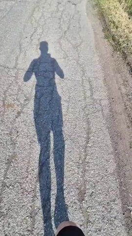 A person's shadow is visible on a cracked asphalt road running through a rural landscape on a sunny day with scattered clouds. Tall grass grows along the roadside. In the distance, low hills and a few trees dot the horizon. A power line pole stands in the field. The camera appears to be held by the person whose shadow is visible.