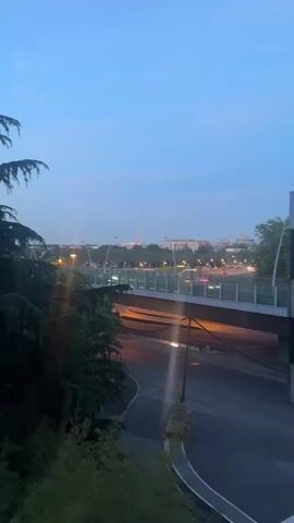 The video, shot from an elevated perspective, presents an urban landscape at dusk. In the foreground, lush green trees partially obscure the view, while a highway overpass dominates the midground. Cars and trucks speed along the road beneath the overpass, their headlights casting streaks of light. In the distance, the Eiffel Tower is visible, its lights illuminating the Paris skyline against a darkening blue sky. A DHL truck passes in front of the camera briefly obscuring the view.