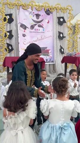 This media file captures a graduation ceremony for young children at "Snow White Modern School" in Gaza. The scene is indoors, with white draped fabric overhead and a stage area with a glass floor. The backdrop features a banner with the school's name, a congratulatory message, and graduation-themed graphics like mortarboards and books. Several children in formal attire, including dresses and shirts, are present.

A man in a green garment and headscarf is leading the ceremony, gesturing and engaging with the children. Another woman in a black hijab and a maroon dress stands to his side. The children appear excited and are clapping, some smiling broadly. A girl in a white dress and another in a light blue dress are prominently featured, facing away from the camera. The overall atmosphere is celebratory and joyful. The visible text on the banner reads "S.M.S Snow White Modern School" and "Congratulations" with percentages indicating high achievement. The lighting suggests it is daytime.