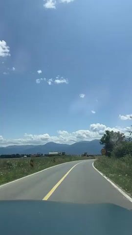 A car is driving on a paved road with a yellow line down the middle. The road is surrounded by green grass and fields. In the distance, there are mountains. The sky is blue with white clouds. There is a speed limit sign for 40 on the right side of the road. There are some construction vehicles in the distance on the left side of the road. The sun is shining.