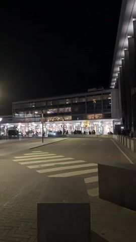 The video shows an outdoor scene at night. The surroundings are dark, illuminated by street lights and building lights. There's a paved area, possibly a plaza or walkway, made of rectangular blocks. There are crosswalks painted with white stripes. A modern building with a glass facade and interior lighting can be seen in the background. Vehicles, including taxis (one is yellow), are parked and driving in the distance. There's a bus shelter made of glass and metal. A billboard with text and an image is also visible in the background. The overall scene looks like an airport or transportation hub at night.