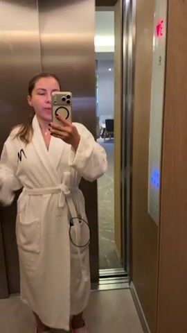 A woman is standing in an elevator, taking a selfie. She is wearing a white bathrobe and pink Crocs. She is holding a water bottle in her left hand and her phone in her right hand. The elevator doors are made of stainless steel and there is a panel of buttons on the right side of the elevator. The woman's hair is pulled back in a ponytail. She has on black nail polish. The woman appears to be in her twenties or thirties. She is making a pouty face at the camera.