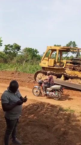 The video shows an outdoor scene featuring a dirt road and several individuals. In the foreground, a man wearing a grey polo shirt and dark trousers stands, looking down at his smartphone. He has a sturdy build. Behind him, another man is seen riding a black motorcycle along the reddish-brown dirt path. To the far right, a large yellow bulldozer is partially visible, its tracks on the ground, and the text "D-8K" can be seen on its side.

The setting appears to be a rural or semi-rural area, likely in Otunade, Nigeria, given the context. The ground consists of loose, reddish-brown earth, suggesting recent clearing or road construction. Lush green trees and dense foliage line both sides of the dirt path and extend into the background under a bright, clear sky. The sunlight indicates it is daytime, possibly midday or early afternoon. The overall impression is one of development or infrastructure work in a natural, vegetated environment.