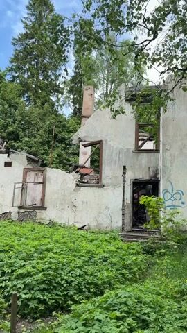 The video captures a wide shot of a ruined, abandoned building, likely a former house, set amidst lush greenery in Tokkari-Lend, Russia. The structure exhibits extensive damage, with charred wooden beams and roof sections indicating it was likely destroyed by fire. Many window frames are empty or contain broken panes, and portions of the light-colored walls appear crumbled and distressed. A brick chimney stands partially intact on the right side of the building.

The foreground is covered with a dense growth of vibrant green plants, suggesting the area is overgrown and unmaintained. Behind and surrounding the ruined building, tall, mature trees, a mix of coniferous and deciduous, form a dense wooded backdrop. The sky above is blue with scattered white clouds, indicating a bright, clear daytime. A small, dark blue information sign on a stake is visible in the lower left foreground, though its text is not legible. The scene evokes a sense of abandonment and the forces of nature reclaiming a once-inhabited space.