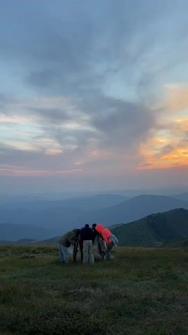 The video captures a group of people gathered on a grassy mountaintop, looking down at something on the ground. They are silhouetted against a vast mountain landscape under a cloudy sky. The sun is setting, casting an orange and pink glow across the horizon. The mountains are layered in shades of blue and gray, creating a sense of depth. The overall mood is serene and contemplative.
