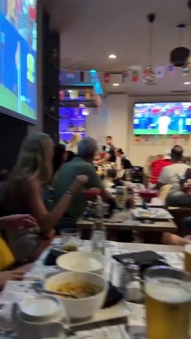 A group of people are watching a soccer game on a large television screen in a dimly lit bar. The screen displays a soccer player in a white uniform with the number six on his back, raising his arms in celebration. Several other players in white uniforms are also visible on the field. The bar is decorated with small flags from various countries. In the foreground, tables are set with food and drinks. A woman with long blonde hair is seated on the left, facing the screen. A man with gray hair is seated next to her, also facing the screen. Other patrons are seated at tables, watching the game. A person in a black t-shirt and shorts is standing in front of the bar, appearing to interact with the patrons. A projector screen on the left also shows a soccer game.