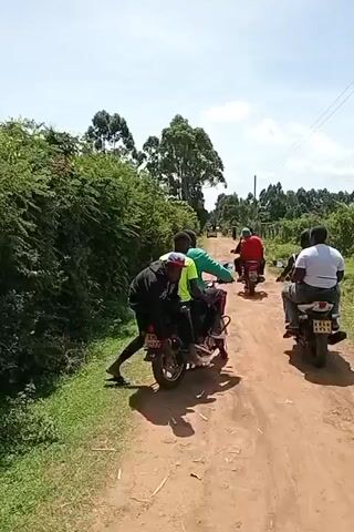 Here's a detailed description of the video:

The video shows a group of people riding motorcycles on a dirt road through a rural area. The camera is positioned on one of the motorcycles, facing forward, capturing the perspective of a rider or passenger.

Several motorcycles are ahead on the dirt road. Most of the motorcycles appear to have two riders each. The riders and passengers are all young men, dressed in casual clothing. The first motorcycle has three people on it; the person in the rear is pushing with their foot to get started.

The environment consists of a dirt road bordered by lush green vegetation, including trees and bushes. The sky is bright, suggesting a sunny day. In the distance, more trees and vegetation are visible, giving the impression of a remote location.

The motorcycles are moving along the road, kicking up dust as they travel. The riders appear to be enjoying the ride.