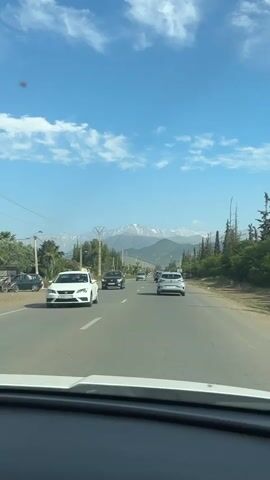 The video shows a car driving on a road with a view of mountains in the distance. The sky is blue with scattered clouds. There are other cars and motorcycles on the road. The road is lined with trees and utility poles. The mountains in the background have snow-capped peaks. The view is from the perspective of the driver, looking through the windshield.