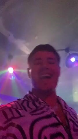 The video shows a group of people dancing in a dimly lit club. The lighting is colorful, with purple and green lights illuminating the scene. One person in the foreground, wearing a black and white striped shirt, is looking at the camera and appears to be singing or talking. Another person, wearing a black cap, is partially visible to their right. Other people are seen dancing in the background.