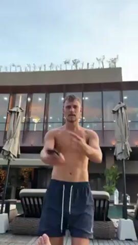 A shirtless man wearing dark blue shorts is standing outdoors, possibly near a pool, and is doing arm movements, stretching or lightly dancing.  Behind him is a building with multiple glass panels and a rooftop with what appears to be small trees or shrubbery.  There are lounge chairs with cream colored cushions and draped sun umbrellas in the background. The sky is light, suggesting it is late afternoon or early evening.