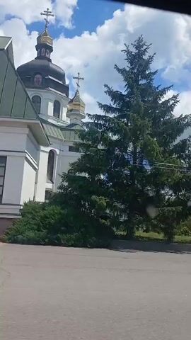 The video shows a moving camera panning from right to left, capturing a street scene with a church and trees. The church has a dark dome with a gold cross on top, and its white walls are visible. Tall green pine trees are in the foreground, obscuring some of the view. In the background, there are other buildings and a clear blue sky with white clouds. The road in front is paved.