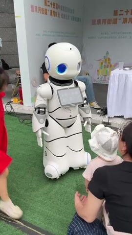 A white humanoid robot with blue glowing eyes stands on a green surface resembling artificial turf. It has a tablet mounted on its chest. A young girl in a red dress with a red hair accessory is interacting with the robot. She extends her arm toward the robot's chest. To the right of the girl, partially obscured, are two people, one wearing a white hat. In the background, a man sits in a chair. Behind the robot and people is a white wall with text and graphics, suggesting an event or exhibition. The robot's screen is displaying a gray interface.