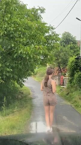 A lone female figure, viewed from behind, walks away from the camera on a wet asphalt road. She has long brown hair tied in a ponytail and is wearing a matching light brown sleeveless top and shorts set with white sneakers. The road is bordered by dense green foliage, including tall trees on the left and bushes on the right. A pile of stacked firewood is visible behind the bushes on the right. Utility poles and wires are present in the distant background. The ground is visibly wet, reflecting light, indicating recent rain. The camera appears to be inside a vehicle, with a slight reflection visible at the bottom of the frame.