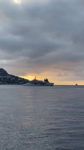 The video shows a body of water with several large yachts. The sky is overcast with some patches of sunlight breaking through. There are hills with buildings visible in the background. The camera pans across the scene, showing different views of the yachts and the coastline. The water is choppy, suggesting a moderate breeze. There are no people visible in the video.