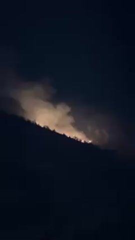 Here is a description of the video:

The video shows a wildfire burning on the side of a hill at night. The fire is visible as a bright orange glow, and thick smoke billows into the air.  A person is briefly visible at the bottom of the frame. The rest of the scene is dark, with only the fire illuminating the landscape. The person in the video says "Allahu Akbar".