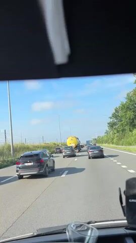 The video shows a view from inside a vehicle on a highway. Multiple cars and a yellow tanker truck are visible on the road ahead, suggesting traffic congestion. The sky is blue with some clouds, and trees line the right side of the highway. The interior of the vehicle, including the dashboard and windshield, is partially visible in the foreground.