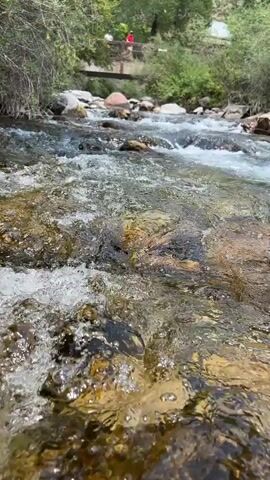 The video shows a close-up, low-angle shot of a clear, shallow stream flowing over a rocky bed. The water is in motion, creating ripples and small splashes in the foreground. Sunlight illuminates the water, revealing the multicolored rocks beneath the surface. In the background, a wooden bridge crosses the stream, and two figures, one wearing a red shirt, are visible standing on the bridge. Lush green vegetation lines the banks of the stream. The overall scene depicts a natural, outdoor environment with a flowing body of water.