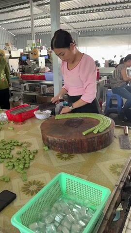 In an indoor workshop in Thới Sơn, Vietnam, a woman wearing a pink long-sleeved shirt, a black apron, and a smartwatch is meticulously slicing long, green confectionery on a large, thick wooden chopping block. She uses a cleaver-style knife to cut the elongated green pieces, possibly a type of mung bean cake, into smaller, uniform cubes. Several cut pieces are already scattered across the patterned table next to the chopping block. In the foreground, a green plastic basket holds numerous small, clear plastic containers, indicating that the cut sweets are being prepared for packaging. Another woman is visible in the background, seated and working, surrounded by industrial equipment and shelving, suggesting a small-scale production or packaging facility. The scene captures a precise step in the preparation of traditional Vietnamese sweets.