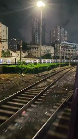 The video is shot from a moving train at night. The camera is pointed forward and slightly to the right, showing train tracks stretching into the distance. To the left of the tracks, there is a row of parked trains with their interior lights on, suggesting they are at a station or railyard. Beyond the trains, there are buildings, including a tall, unfinished skyscraper under construction and several apartment buildings with illuminated windows. Overhead power lines for the trains are visible, supported by metal poles. The ground around the tracks is covered in gravel and debris. The overall atmosphere is urban and industrial, illuminated by the artificial lights of the train, buildings, and streetlights.