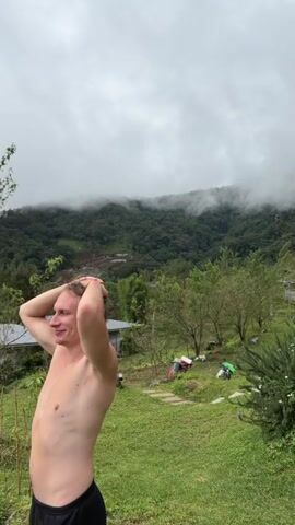 A young man with blond hair is standing outdoors with his hands on his head. He is facing left and looking slightly up and to his left. He is wearing black shorts and is shirtless. Behind him, there is a grassy yard with trees, some houses with white roofs, and a misty, tree-covered mountain in the background. The sky is overcast.