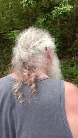 A person, viewed from behind, walks away from the camera on what appears to be a dirt or gravel path. They have long, curly, gray hair tied back with a green hair tie, and a long, full gray beard. They are wearing a dark gray tank top. Lush green trees and foliage form the background, suggesting an outdoor, possibly wooded, setting. An off-camera voice asks if something is "freaking you out" and mentions "he's looking at me like that," implying an interaction with an unseen entity, possibly an animal. The person continues to walk forward.