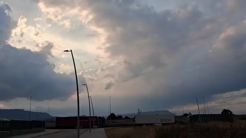 The video captures a wide shot of an outdoor scene, possibly an industrial or logistical area, under a dramatic sky. The camera pans slowly from left to right, revealing a road, a sidewalk, and a metal fence in the foreground. Behind the fence, several large trucks are visible, some with red trailers. In the background, mountains can be seen, with hints of buildings on the mountaintops.

The sky is filled with large, dark clouds, suggesting impending rain or a storm. However, shafts of sunlight are breaking through the clouds, creating a theatrical and somewhat ominous atmosphere. The light is warm and golden in places, indicating it is likely late afternoon or early evening. The overall mood is one of quiet observation, with the vastness of the sky and the stoic presence of the trucks and mountains dominating the view.

No people are visible in the media. The main objects of focus are the trucks, the road infrastructure, and the natural landscape. The setting appears to be a functional area, perhaps a truck stop or distribution center, given the presence of multiple large vehicles. The distant mountains and the cloudy sky provide a sense of place, hinting at a location in a region with rolling hills or mountains.

One truck trailer has visible text: "Botyr". The time of day is strongly suggested by the quality of light filtering through the clouds. The weather is clearly overcast with breaks of sunshine. The location cues point towards Gurb, Spain, as indicated by the prompt. There are no overt displays of emotion, but the dramatic sky could evoke feelings of awe or apprehension.