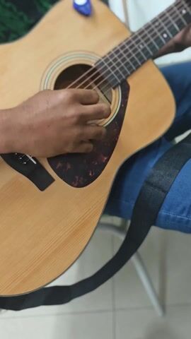 A close-up shot shows a person playing an acoustic guitar. The individual, wearing blue jeans, is sitting down, with the guitar resting on their lap. Their left hand is fretting the strings while their right hand strums the strings near the sound hole, with a guitar pick. A black guitar strap is visible, slung across their body. A blue guitar pick is clipped to the headstock of the guitar. The guitar has a light brown finish with a dark tortoiseshell pickguard. The background is slightly blurred, showing glimpses of a room with light-colored walls, a green corner, and a white object behind the player. The person is playing the guitar with focus.